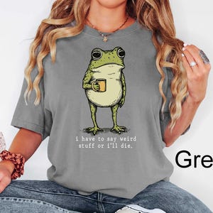 May include: Grey t-shirt featuring a cartoon frog holding a mug. The frog is green and beige, with the text "i have to say weird stuff or i'll die." printed below the frog. The word "Grey" is printed in black on the bottom right.