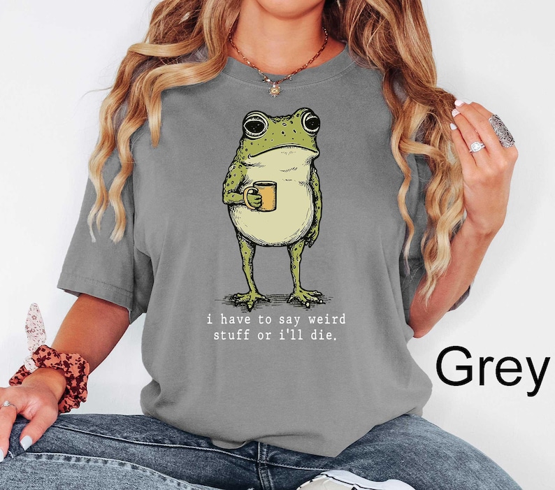 May include: Grey t-shirt featuring a cartoon frog holding a mug. The frog is green and beige, with the text "i have to say weird stuff or i'll die." printed below the frog. The word "Grey" is printed in black on the bottom right.