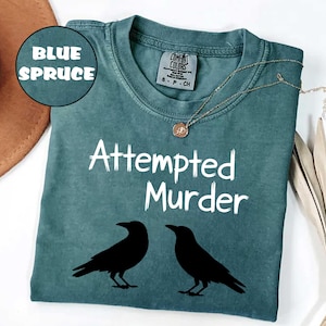 May include: Blue spruce-colored t-shirt with the text "Attempted Murder" and two black crow silhouettes. The shirt has a crew neck and short sleeves. A gold-colored necklace with a pendant is draped on the shirt.