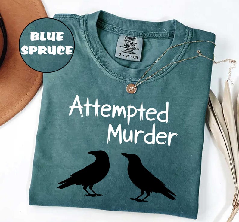 May include: Blue spruce-colored t-shirt with the text "Attempted Murder" and two black crow silhouettes. The shirt has a crew neck and short sleeves. A gold-colored necklace with a pendant is draped on the shirt.