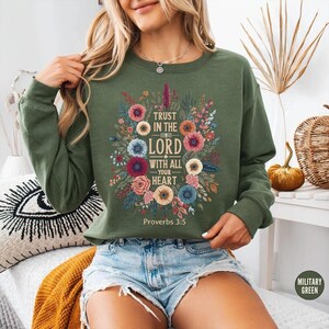 Peut inclure: Sweat-shirt vert militaire avec un motif floral et le texte "Trust in the Lord with all your heart" et "Proverbes 3:5". Le motif floral comprend des fleurs roses, bleues et jaunes.