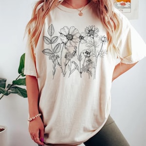May include: Cream-colored t-shirt featuring a black floral line drawing across the chest. The design includes various flowers and foliage. The shirt is a relaxed fit, with rolled-up sleeves. The model is wearing dark green shorts.