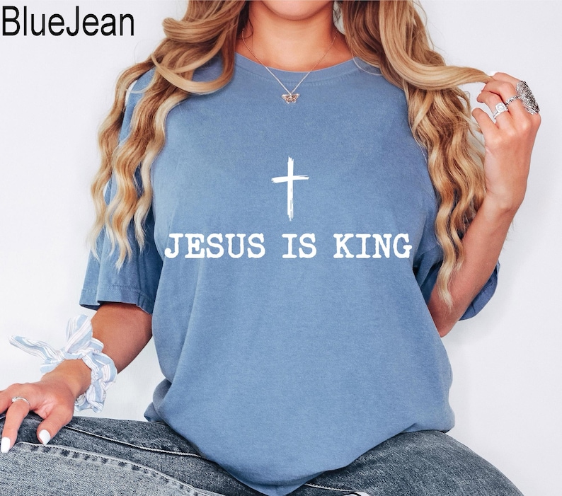 K&ouml;nnte beinhalten: Hellblaues T-Shirt mit dem wei&szlig;en Text "JESUS IS KING" und einem wei&szlig;en Kreuz. Das Shirt hat kurze &Auml;rmel und einen Rundhalsausschnitt. Der Markenname "BlueJean" befindet sich oben links.