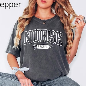 Peut inclure: Un t-shirt gris foncé avec le mot "NURSE" en lettres blanches. En dessous, le nom "RACHEL" est inscrit dans un rectangle blanc, avec un graphique de stéthoscope. Le t-shirt est de coupe ample et confortable.