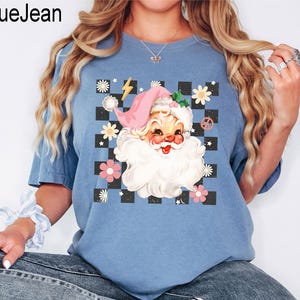 May include: Blue t-shirt with a Santa Claus graphic. The Santa Claus illustration features a pink hat, white beard, and a checkerboard background with flowers and a peace sign. The text "BlueJean" is in the upper left corner.