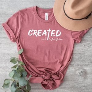 May include: A maroon t-shirt with white text that reads "CREATED with a purpose" and a white cross.