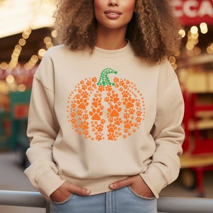 Peut inclure: Sweat-shirt beige avec un motif citrouille en empreintes de pattes orange et une tige verte. La citrouille est compos&eacute;e de nombreuses empreintes de pattes orange. Un haut d&eacute;contract&eacute; pour l'automne et Halloween.