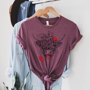 May include: A maroon t-shirt with a black and white illustration of a Highland cow wearing a red bandana and holding a red flower in its mouth.
