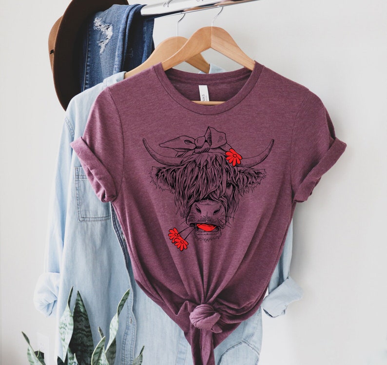 May include: A maroon t-shirt with a black and white illustration of a Highland cow wearing a red bandana and holding a red flower in its mouth.