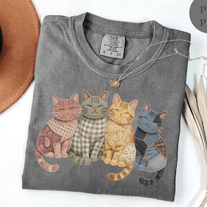 May include: A gray Comfort Colors t-shirt featuring four smiling cat appliqués with patchwork patterns in shades of pink, gray, yellow, and blue. The shirt has a crew neck and a small tag at the neckline.
