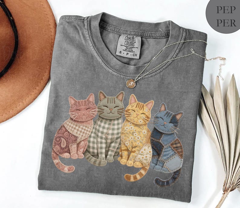 May include: A gray Comfort Colors t-shirt featuring four smiling cat appliqu&eacute;s with patchwork patterns in shades of pink, gray, yellow, and blue. The shirt has a crew neck and a small tag at the neckline.