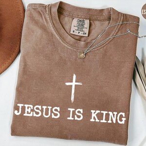 K&ouml;nnte beinhalten: Espressofarbenes T-Shirt mit dem Text "JESUS IS KING" und einer wei&szlig;en Kreuzgrafik. Das Shirt hat einen Rundhalsausschnitt und kurze &Auml;rmel. Eine silberne Halskette mit Anh&auml;nger liegt auf dem Shirt.