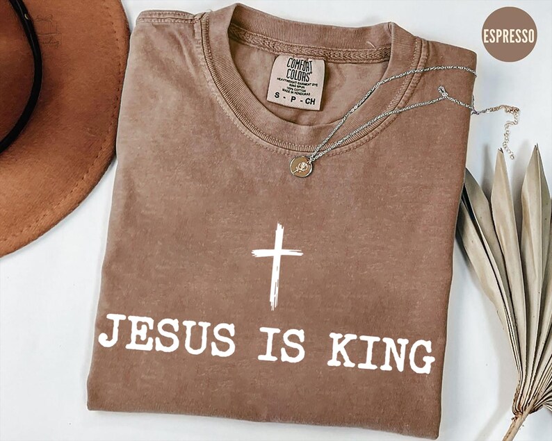 K&ouml;nnte beinhalten: Espressofarbenes T-Shirt mit dem Text "JESUS IS KING" und einer wei&szlig;en Kreuzgrafik. Das Shirt hat einen Rundhalsausschnitt und kurze &Auml;rmel. Eine silberne Halskette mit Anh&auml;nger liegt auf dem Shirt.