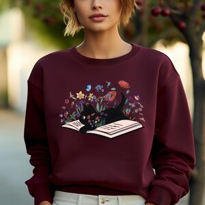 May include: A maroon sweatshirt with a black cat reading a book surrounded by flowers. The cat is lying on the open book, which is surrounded by a variety of flowers in shades of pink, purple, blue, yellow, and green.