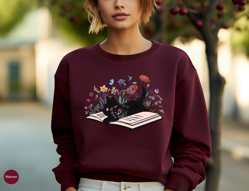 May include: A maroon sweatshirt with a black cat reading a book surrounded by flowers. The cat is lying on the open book, which is surrounded by a variety of flowers in shades of pink, purple, blue, yellow, and green.