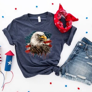 May include: A navy blue t-shirt with a graphic of an American bald eagle with an American flag design. The eagle is in front of a red, white, and blue splattered background.
