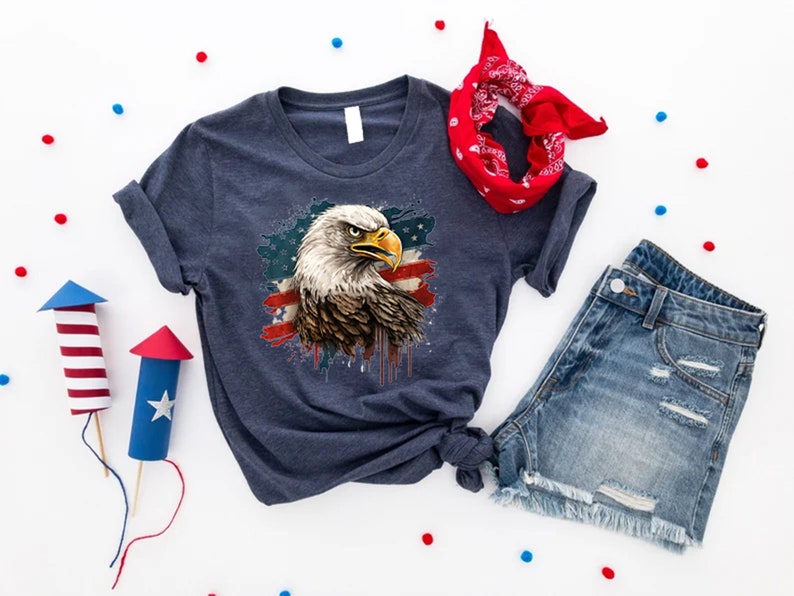 May include: A navy blue t-shirt with a graphic of an American bald eagle with an American flag design. The eagle is in front of a red, white, and blue splattered background.