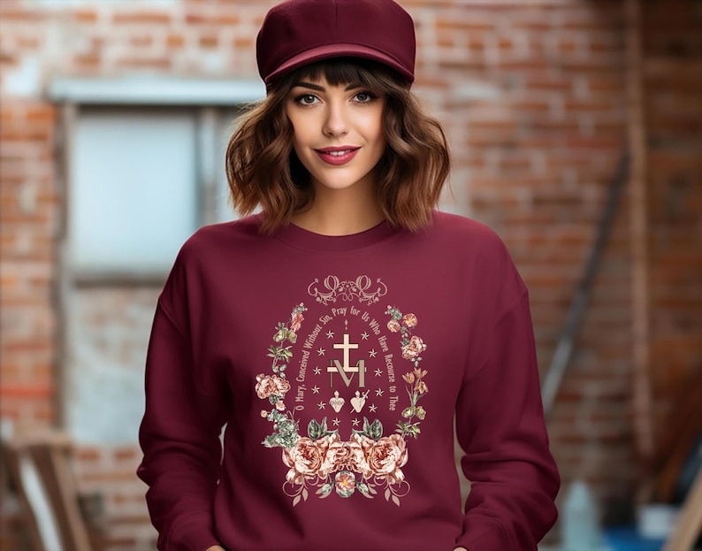 Puede incluir: Una sudadera color burdeos con un dise&ntilde;o floral y el texto "Reza por nosotros que recurrimos a Ti" en un c&iacute;rculo alrededor de una cruz y las letras "M" y "A".