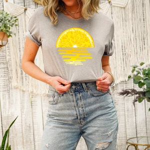 Pu&ograve; includere: Una maglietta grigia con una grafica di una fetta di limone gialla con un tramonto alle spalle.