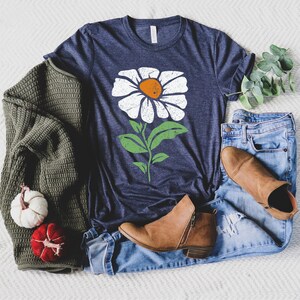 May include: A navy blue t-shirt with a white daisy flower graphic. The flower has a green stem and leaves. The t-shirt is laying on a white surface with a green sweater, blue jeans, and brown boots.