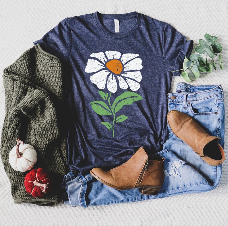May include: A navy blue t-shirt with a white daisy flower graphic. The flower has a green stem and leaves. The t-shirt is laying on a white surface with a green sweater, blue jeans, and brown boots.