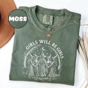K&ouml;nnte beinhalten: Ein moosgr&uuml;nes T-Shirt mit dem Text "GIRLS WILL BE GIRLS" und einer Illustration von f&uuml;nf Hexen. Das Shirt hat einen Rundhalsausschnitt und kurze &Auml;rmel. Eine goldene Halskette mit Anh&auml;nger liegt &uuml;ber dem Shirt.