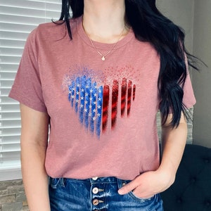 Pu&ograve; includere: Una maglietta color malva con un disegno a forma di cuore. Il disegno incorpora elementi della bandiera americana, con stelle blu e bianche a sinistra e strisce rosse a destra. La maglietta ha maniche corte e scollo tondo.