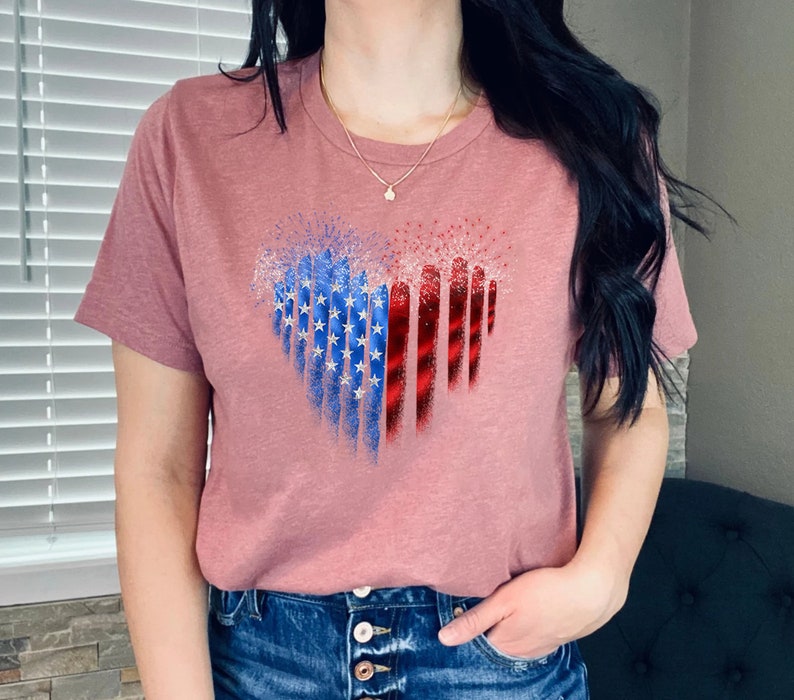 Pu&ograve; includere: Una maglietta color malva con un disegno a forma di cuore. Il disegno incorpora elementi della bandiera americana, con stelle blu e bianche a sinistra e strisce rosse a destra. La maglietta ha maniche corte e scollo tondo.