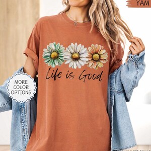 May include: A terracotta-colored t-shirt featuring three daisy flowers in green, white, and orange, with the phrase "Life is Good" in black script. The shirt has rolled-up sleeves and a relaxed fit. A denim jacket is worn over the shirt.