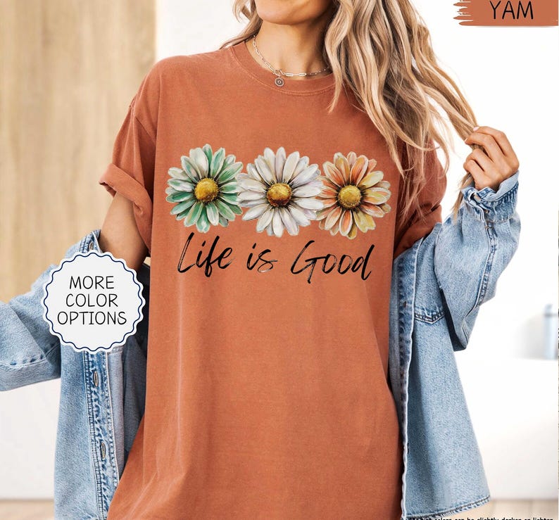 May include: A terracotta-colored t-shirt featuring three daisy flowers in green, white, and orange, with the phrase "Life is Good" in black script. The shirt has rolled-up sleeves and a relaxed fit. A denim jacket is worn over the shirt.