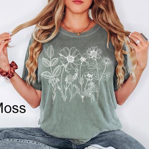 May include: A moss green t-shirt featuring a white floral line drawing design. The word "Moss" is printed in black on the lower left side of the image. The shirt is a short-sleeved, crew neck style.