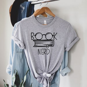 Pu&ograve; includere: Maglietta grigia con una grafica nera di un libro con occhiali in cima e il testo "Book Nerd".