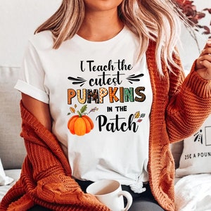 May include: White t-shirt with the text "I Teach the Cutest Pumpkins in the Patch" in black, orange, and green. The shirt features a watercolor pumpkin graphic. The person is wearing an orange cardigan.