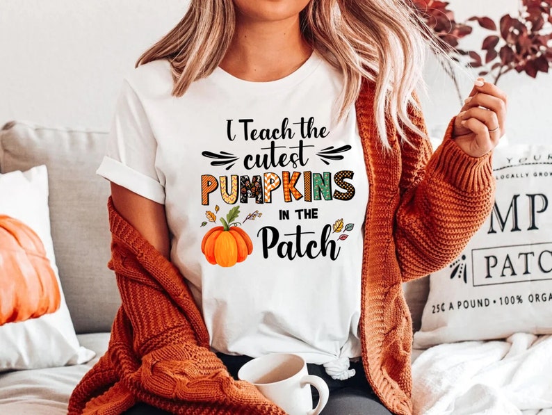 May include: White t-shirt with the text "I Teach the Cutest Pumpkins in the Patch" in black, orange, and green. The shirt features a watercolor pumpkin graphic. The person is wearing an orange cardigan.