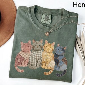 May include: Hemp green t-shirt featuring four smiling cat illustrations with patchwork patterns. The shirt has a crew neck and short sleeves. A silver necklace with a pendant is draped across the shirt. The tag reads "Comfort Colors".