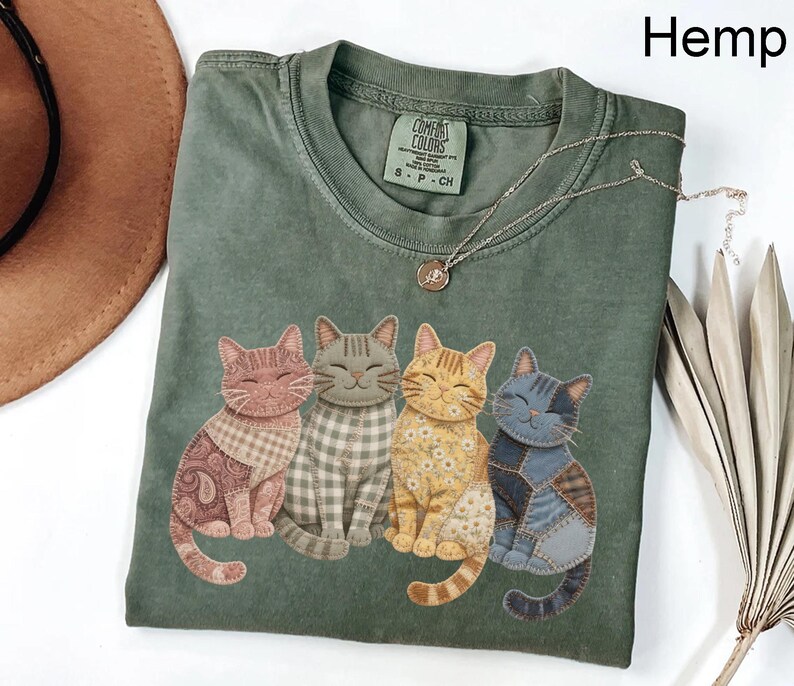 May include: Hemp green t-shirt featuring four smiling cat illustrations with patchwork patterns. The shirt has a crew neck and short sleeves. A silver necklace with a pendant is draped across the shirt. The tag reads "Comfort Colors".