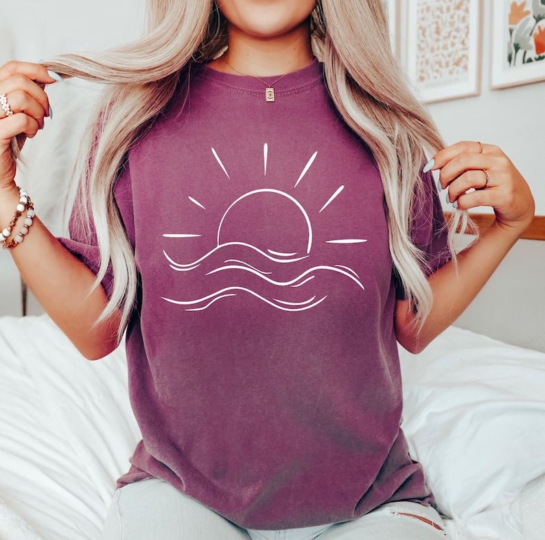 Puede incluir: Camiseta de color ciruela con un dibujo lineal blanco de un sol poni&eacute;ndose sobre las olas del oc&eacute;ano. La camiseta tiene mangas cortas y un corte relajado. El dise&ntilde;o es sencillo y evoca una sensaci&oacute;n de calma. El color es un bonito p&uacute;rpura.
