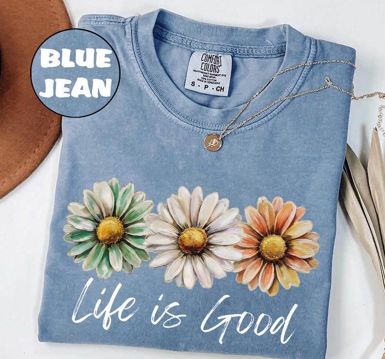 May include: A blue jean Comfort Colors t-shirt with three daisy flowers in green, white, and orange. The words "Life is Good" are printed in white script below the flowers. A gold necklace with a small pendant is draped across the shirt.