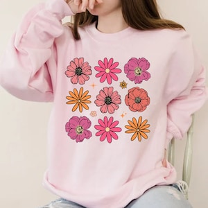Può includere: Una felpa rosa con un design floreale. I fiori sono in tonalità di rosa, arancione e giallo. Ci sono anche piccole stelle sparse in tutto il design.