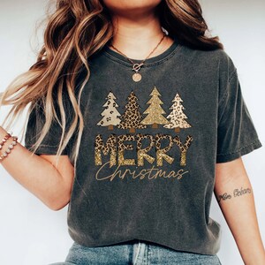 May include: A dark gray t-shirt with a graphic of three Christmas trees decorated with leopard print and gold glitter. The text "Merry Christmas" is printed below the trees in gold glitter.