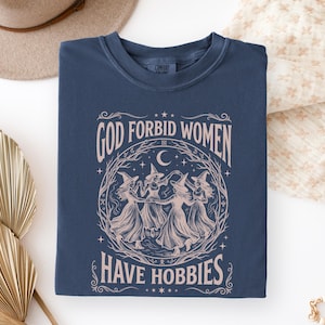 K&ouml;nnte beinhalten: Marineblaues T-Shirt mit dem Text "GOD FORBID WOMEN HAVE HOBBIES" in Rosa. Die Grafik zeigt einen Kreis von Hexen, die unter einem Halbmond tanzen. Das T-Shirt ist gefaltet.
