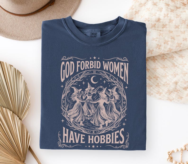 K&ouml;nnte beinhalten: Marineblaues T-Shirt mit dem Text "GOD FORBID WOMEN HAVE HOBBIES" in Rosa. Die Grafik zeigt einen Kreis von Hexen, die unter einem Halbmond tanzen. Das T-Shirt ist gefaltet.