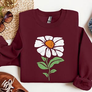 May include: A maroon sweatshirt with a white daisy flower graphic. The flower has a yellow center and green leaves.