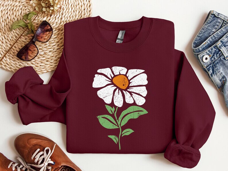 May include: A maroon sweatshirt with a white daisy flower graphic. The flower has a yellow center and green leaves.