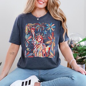 May include: Navy blue t-shirt featuring a colorful illustration of the Statue of Liberty, surrounded by flowers and fireworks. The design incorporates red, yellow, blue, and green hues, creating a vibrant and patriotic aesthetic.