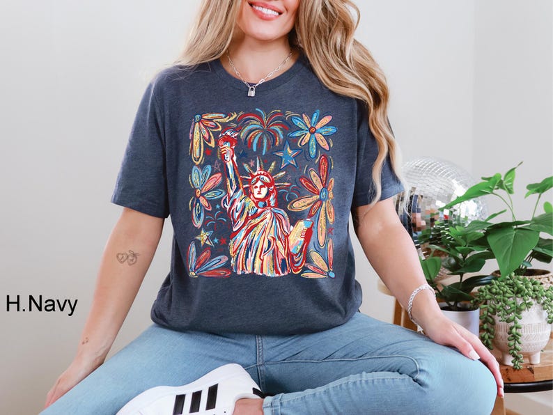 May include: Navy blue t-shirt featuring a colorful illustration of the Statue of Liberty, surrounded by flowers and fireworks. The design incorporates red, yellow, blue, and green hues, creating a vibrant and patriotic aesthetic.