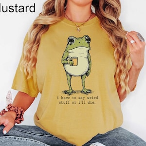 May include: Mustard-colored t-shirt featuring a whimsical illustration of a frog holding a mug. The frog is green with black eyes and is standing upright. The text "i have to say weird stuff or i'll die" is printed below the frog.