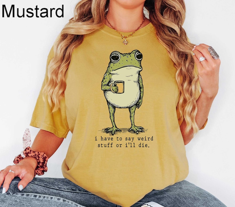May include: Mustard-colored t-shirt featuring a whimsical illustration of a frog holding a mug. The frog is green with black eyes and is standing upright. The text "i have to say weird stuff or i'll die" is printed below the frog.