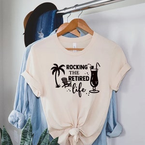Op de afbeelding: Een beige T-shirt met een zwart grafisch ontwerp met de tekst "Rocking the Retired life" met een palmboom, een strandstoel en een cocktailglas.