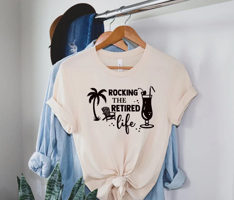 Op de afbeelding: Een beige T-shirt met een zwart grafisch ontwerp met de tekst "Rocking the Retired life" met een palmboom, een strandstoel en een cocktailglas.